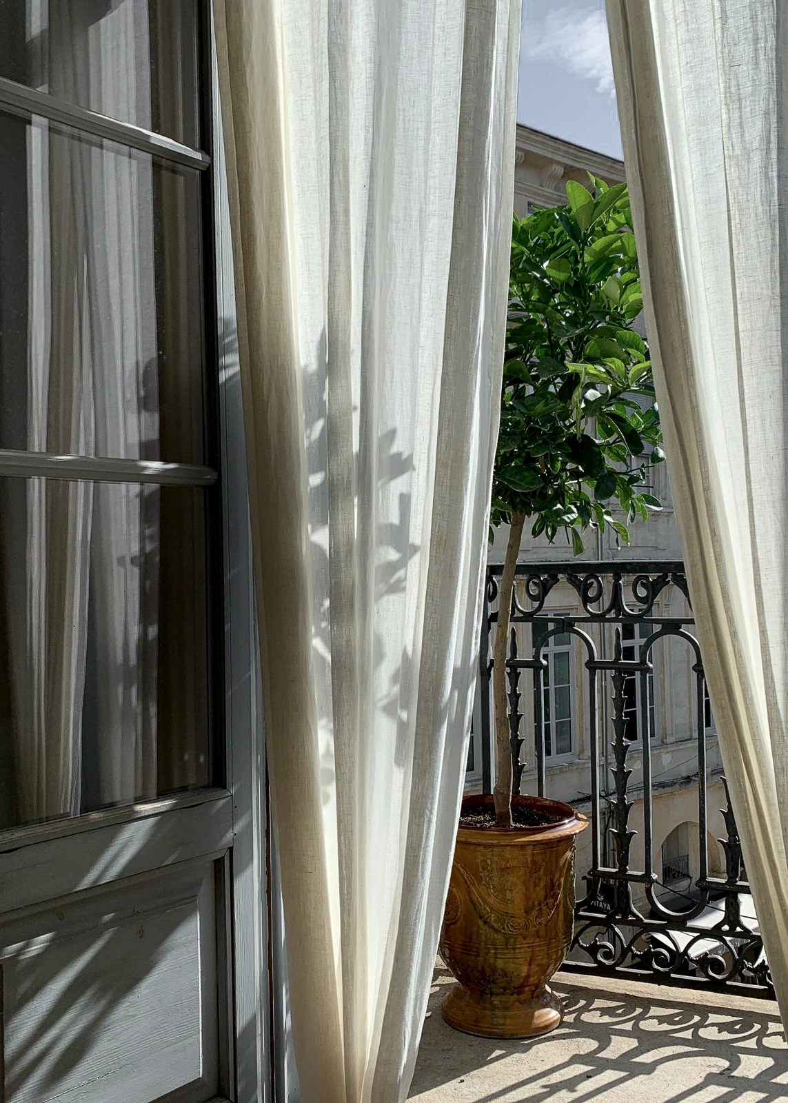 Vue depuis une fenêtre ouverte de l'agence, avec rideaux en lin, balcon ancien et citronnier dans un pot d'Anduze.